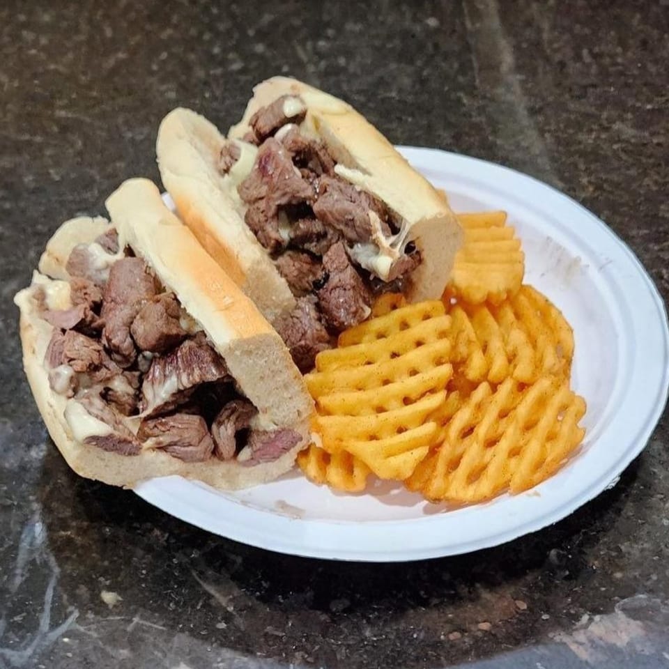 Two cheesesteak sandwiches with waffle fries on a white paper plate from Eastside Market.