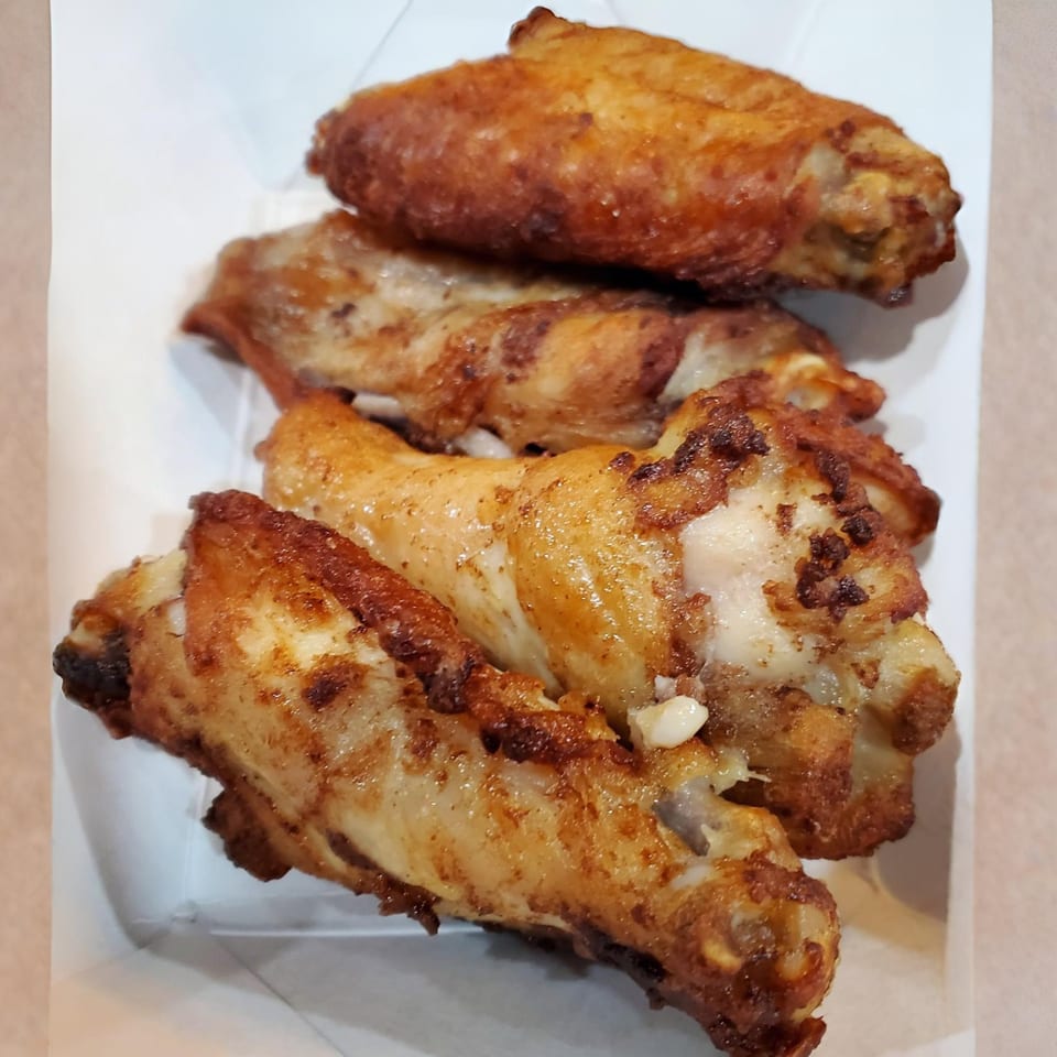 Four crispy, golden-brown chicken wings with visible seasoning, served in a white paper tray.